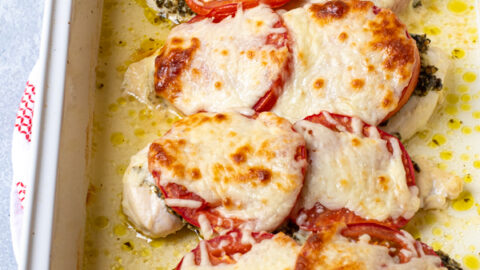 Hands holding a white baking dish containing pesto chicken bake, showing off melted mozzarella cheese on top.