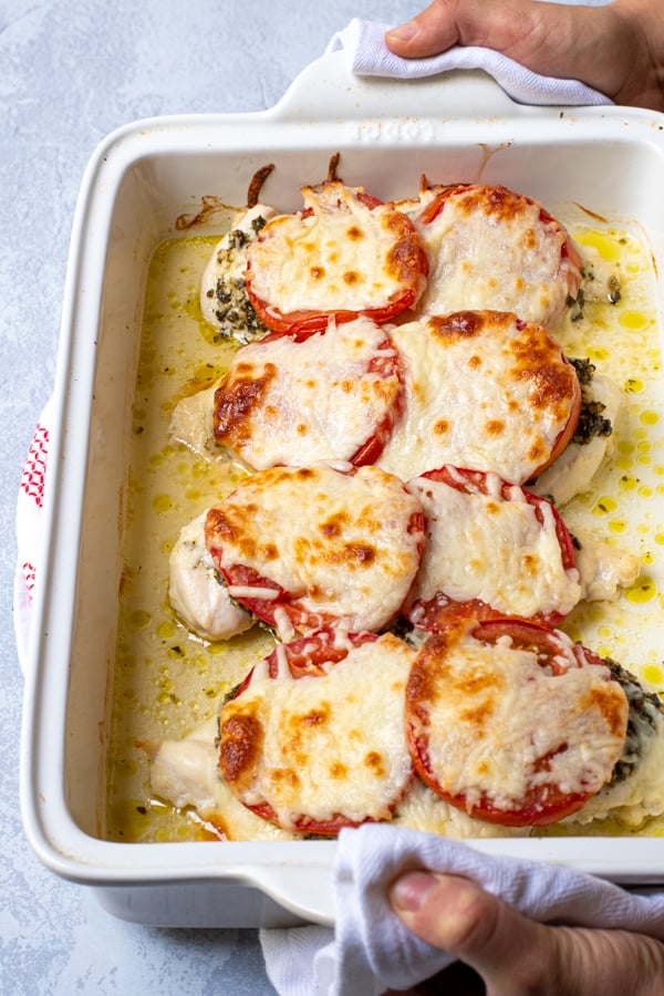 Hands holding a white baking dish containing pesto chicken bake, showing off melted mozzarella cheese on top.