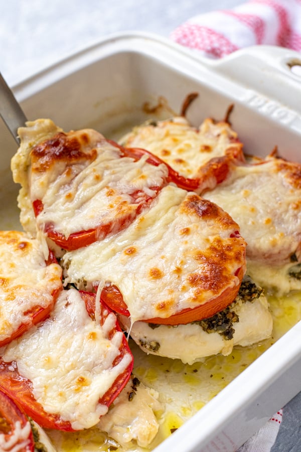 Spatula lifting a baked pesto chicken cutlet out of a dish, covered in melted mozzarella cheese.