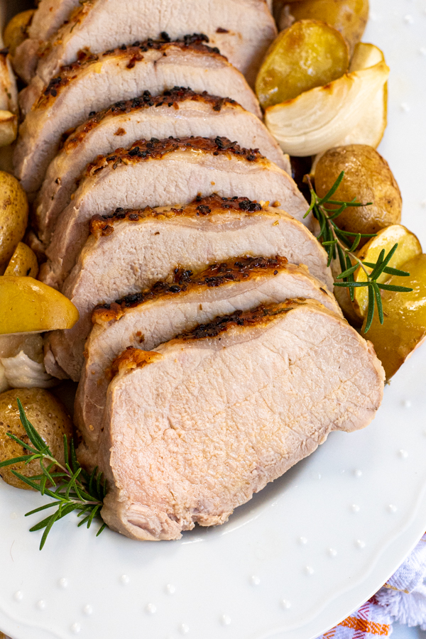 Close up of sliced pork loin on a white serving platter resting next to cut potatoes ad sliced onions and apples.