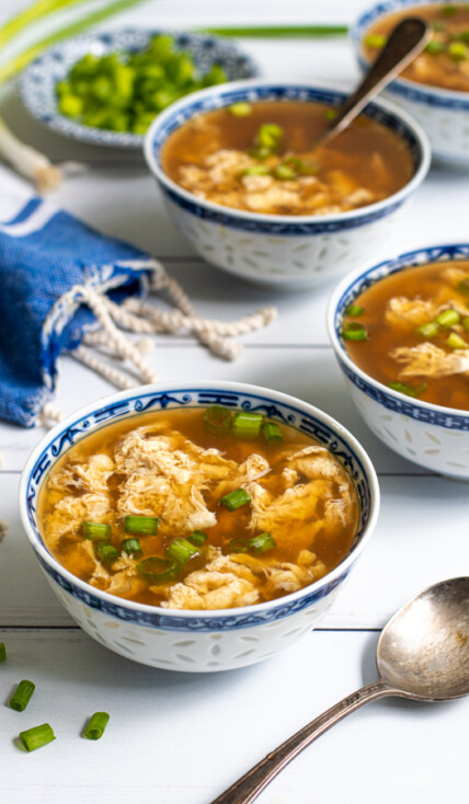Four small Chinese blue and white bowls laid out on a table filled with egg drop soup and garnished with sliced green onions.