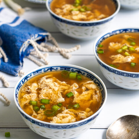 Four small Chinese blue and white bowls laid out on a table filled with egg drop soup and garnished with sliced green onions.