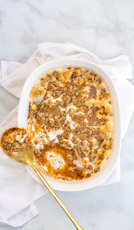Sweet Potato Casserole with Marshmallows and Oat Streusel in a white baking dish with a gold serving spoon sitting on a white kitchen towel.