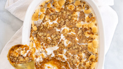 Sweet Potato Casserole with Marshmallows and Oat Streusel in a white baking dish with a gold serving spoon sitting on a white kitchen towel.