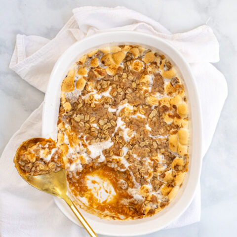 Sweet Potato Casserole with Marshmallows and Oat Streusel in a white baking dish with a gold serving spoon sitting on a white kitchen towel.