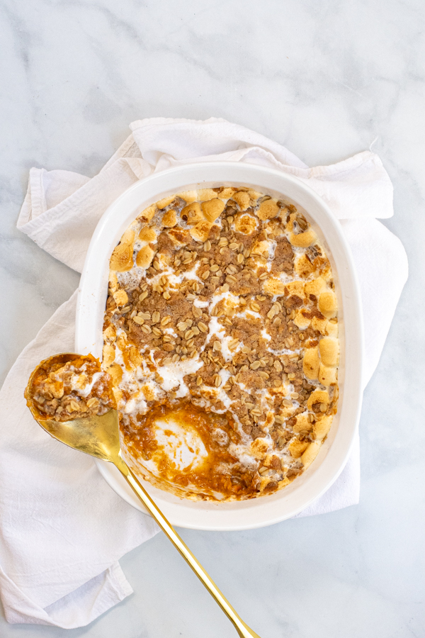 Sweet Potato Casserole with Marshmallows and Oat Streusel in a white baking dish with a gold serving spoon sitting on a white kitchen towel.