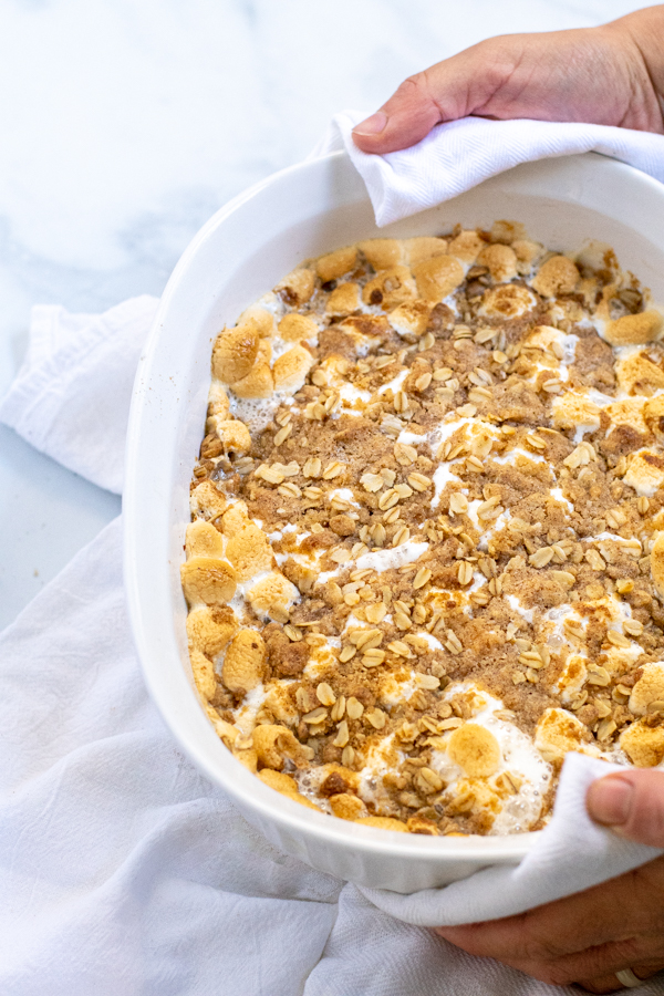 Hands holding a white baking dish with Sweet Potato Casserole with golden Marshmallows and crunchy Oat Streusel.