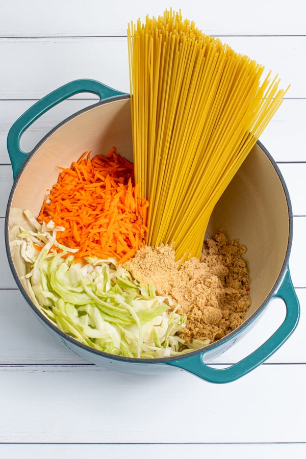 In a large teal dutch oven is uncooked ingredients for teriyaki noodles including spaghetti, cabbage, carrots, and spices.