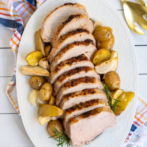 Overhead view of slices of roasted pork loin on a white platter with halved potatoes, apples, and onions.