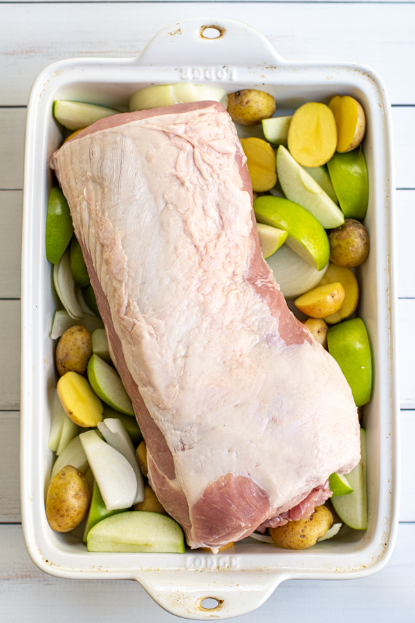 Unseasoned pork loin fat side up sitting in a baking dish with apples, potatoes, and onions.