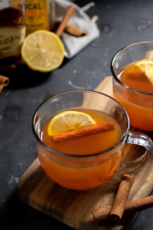 Close up of a clear mug full of hot toddy with a cinnamon stick and a half lemon slice.