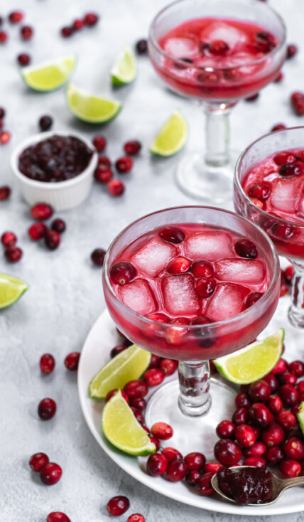 Margarita glasses filled with bright red cranberry sauce margarita and ice, the table that they are on is filled with fresh cranberries and lime slices.