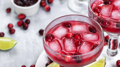Margarita glasses filled with bright red cranberry sauce margarita and ice, the table that they are on is filled with fresh cranberries and lime slices.