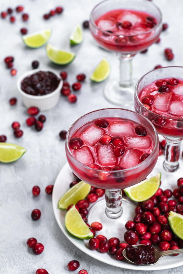 Margarita glasses filled with bright red cranberry sauce margarita and ice, the table that they are on is filled with fresh cranberries and lime slices.