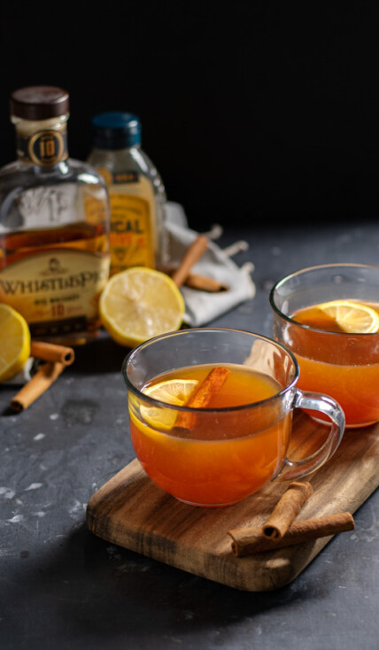 Two clear mugs on a wooden cutting board against a black background filled with hot toddy cocktail and garnished with cinnamon sticks and a lemon wedge.