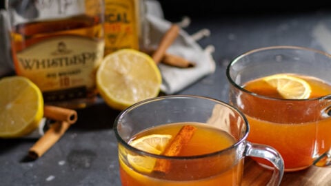 Two clear mugs on a wooden cutting board against a black background filled with hot toddy cocktail and garnished with cinnamon sticks and a lemon wedge.