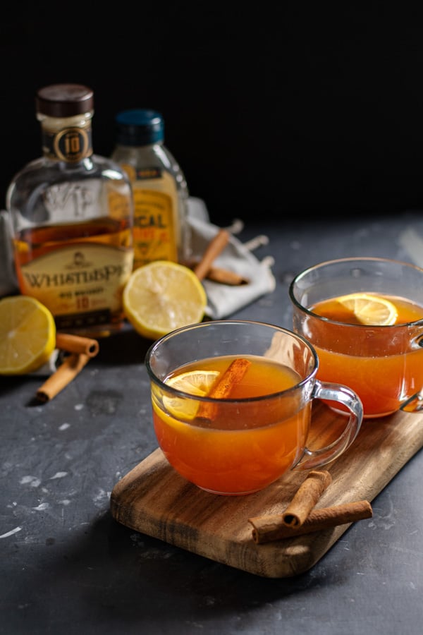 Two clear mugs on a wooden cutting board against a black background filled with hot toddy cocktail and garnished with cinnamon sticks and a lemon wedge.
