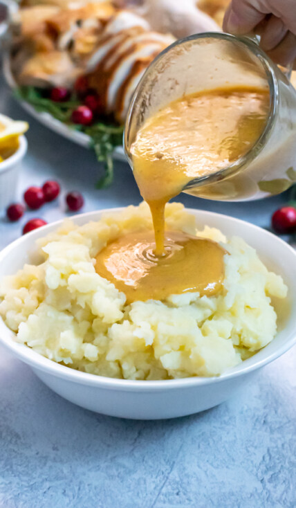 Turkey Gravy From Pan Drippings in a small glass creamer being poured onto a huge bowl of mashed potatoes, the gravy forming a large pool in the center.