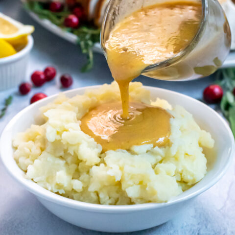Turkey Gravy From Pan Drippings in a small glass creamer being poured onto a huge bowl of mashed potatoes, the gravy forming a large pool in the center.