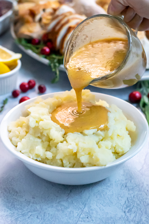 Turkey Gravy From Pan Drippings in a small glass creamer being poured onto a huge bowl of mashed potatoes, the gravy forming a large pool in the center.