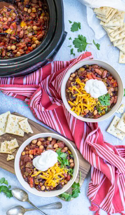 Two bowls and a crockpot full of Three Bean Vegetarian Chili on a table top with garnishes, saltine crackers, a kitchen towels.