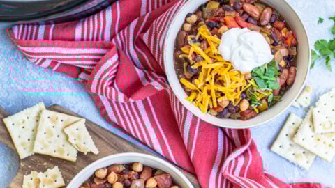 Two bowls and a crockpot full of Three Bean Vegetarian Chili on a table top with garnishes, saltine crackers, a kitchen towels.