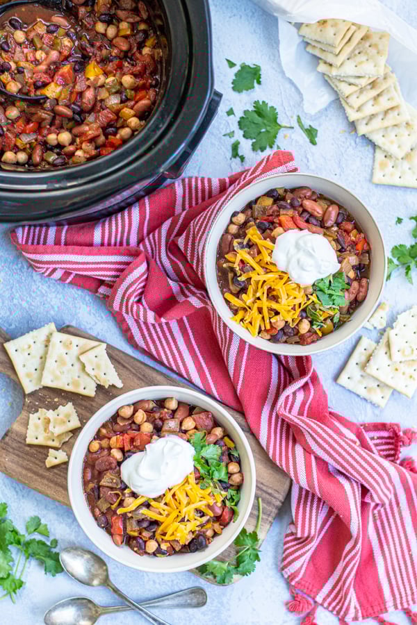 Two bowls and a crockpot full of Three Bean Vegetarian Chili on a table top with garnishes, saltine crackers, a kitchen towels.