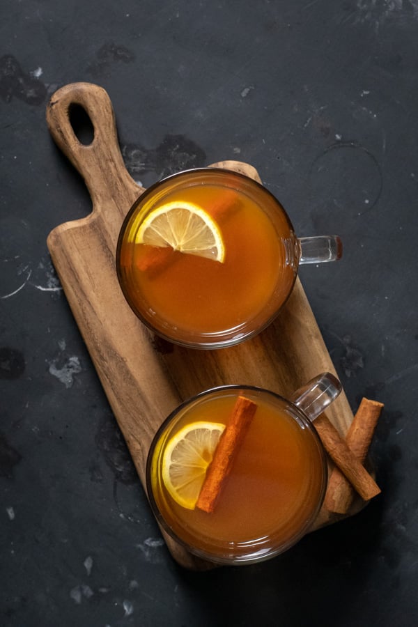 Flatly of a Hot Toddy with Tea Recipe showing two clear mugs filled with a golden liquid and garnished with a cinnamon stick and a lemon slice.