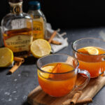 Pinterest Pin with text overlay 'Hot Toddy with Tea, the perfect comforting cocktail'. Image of a clear mug filled with hot toddy and garnished with a cinnamon stick and lemon slice.