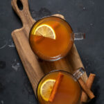 Pinterest Pin with text overlay 'Toddy with Tea, som warm & cozy for winter'. Image of two clear mugs filled with hot toddy and garnished with a cinnamon stick and lemon slice.