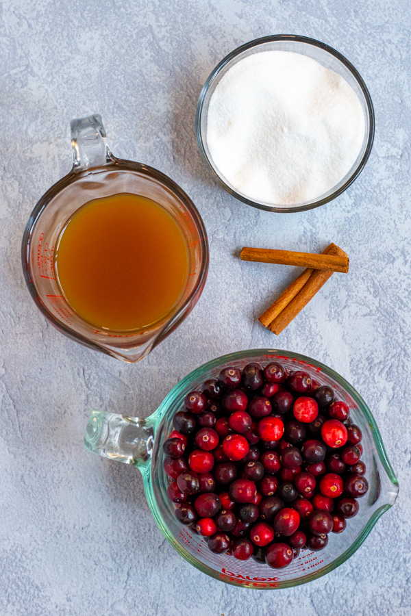Ingredients needed to make cranberry sauce laid on a table including fresh cranberries, apple cider, sugar, and cinnamon sticks.