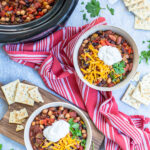 Pinterest Pin with text overlay, The Best Crockpot Vegetarian Chili. Image of two bowls and a crockpot full of chili on a Tavel with spoons, saltine crackers, and a red kitchen towel.