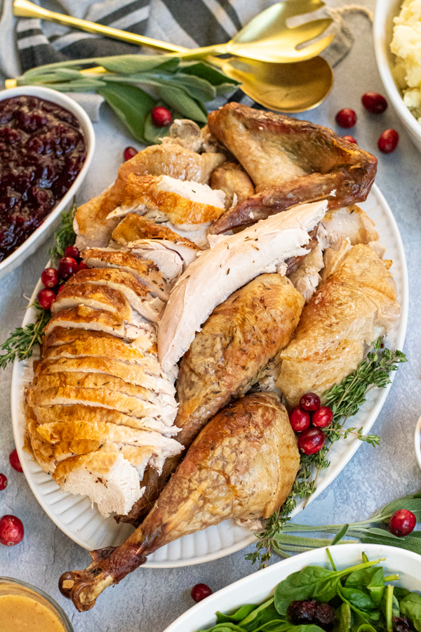 A large white platter with carved dry brined turkey sit-in non a thanksgiving table surrounded by side dishes.