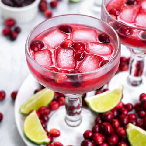 Margarita glass on a white plate filled with cranberries, the glass is filled with a bright red margarita and ice, garnished with more fresh cranberries.