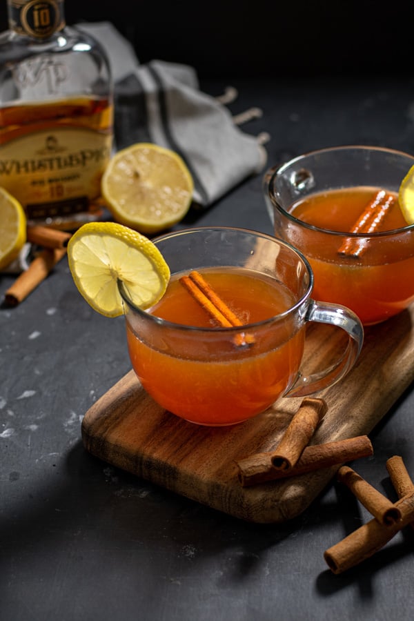 Two clear mugs full of hot toddy on a wooden board against a black background the drinks are garnished with cinnamon sticks and lemon slices.