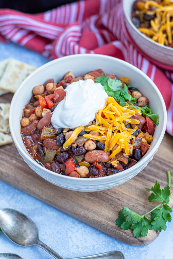A bowl of vegetarian chili garnished with cheddar cheese, sour cream, and cilantro sitting next to some spoons and saltine crackers.