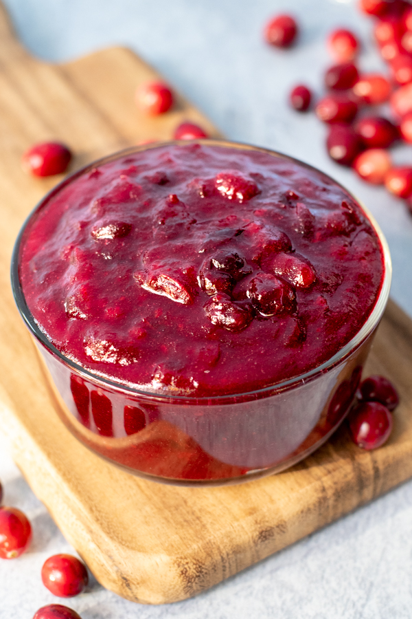 Bowl of cranberry sauce on a wood cutting board with loose fresh cranberries all around.