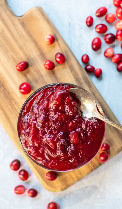 A clear bowl full of bright red homemade cranberry sauce sitting on a wooden cutting board with a spoon in the sauce, next to the cutting board is bunch of loose fresh cranberries.