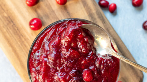 A clear bowl full of bright red homemade cranberry sauce sitting on a wooden cutting board with a spoon in the sauce, next to the cutting board is bunch of loose fresh cranberries.