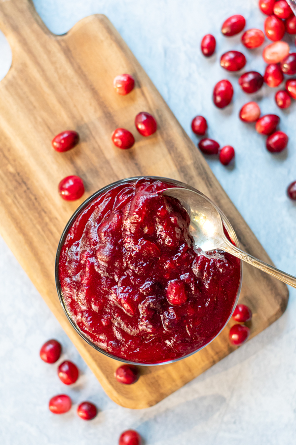 A clear bowl full of bright red homemade cranberry sauce sitting on a wooden cutting board with a spoon in the sauce, next to the cutting board is bunch of loose fresh cranberries.