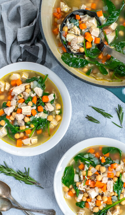 Two bowls and a pot filled with healthy leftover turkey soup on a table next to some spoons for eating and a kitchen towel.