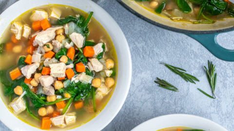 Two bowls and a pot filled with healthy leftover turkey soup on a table next to some spoons for eating and a kitchen towel.