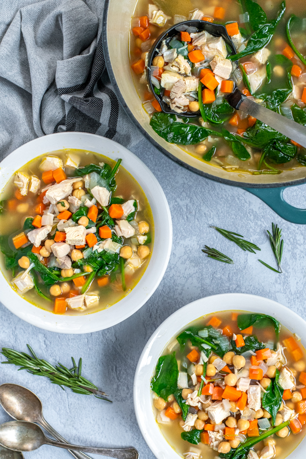 Two bowls and a pot filled with healthy leftover turkey soup on a table next to some spoons for eating and a kitchen towel.
