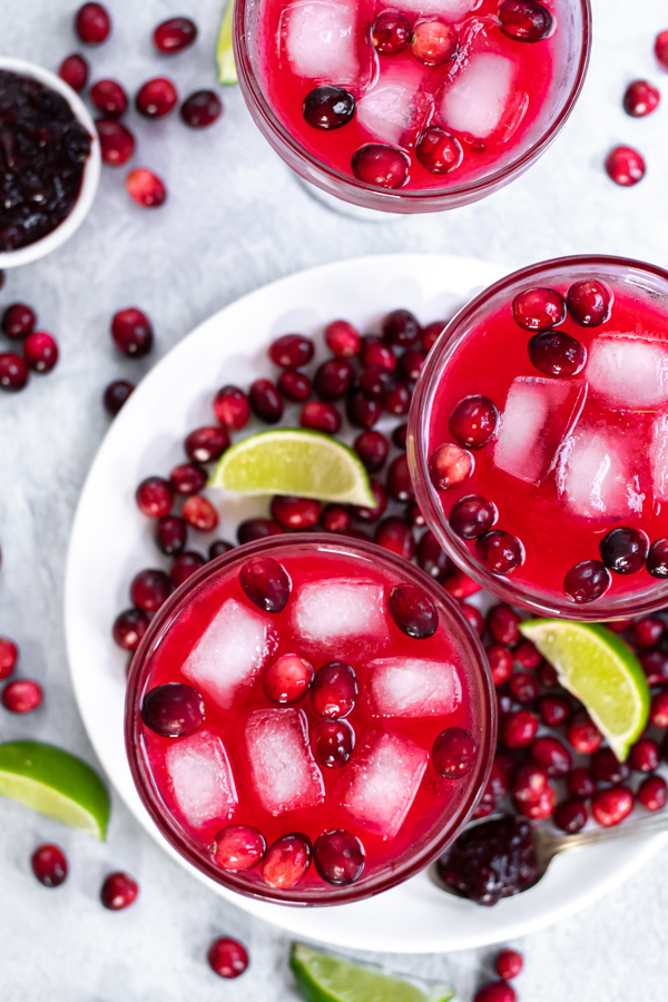 Overhead view of three margarita glasses filled with a bright red drink and ice garnish with fresh cranberries, in addition fresh cranberries are all over the table.