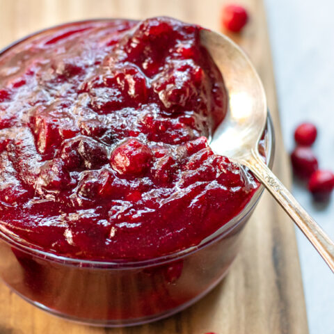 Small bowl of cranberry sauce with a spoon in the bowl sitting on a wooden cutting board next to loose fresh cranberries.