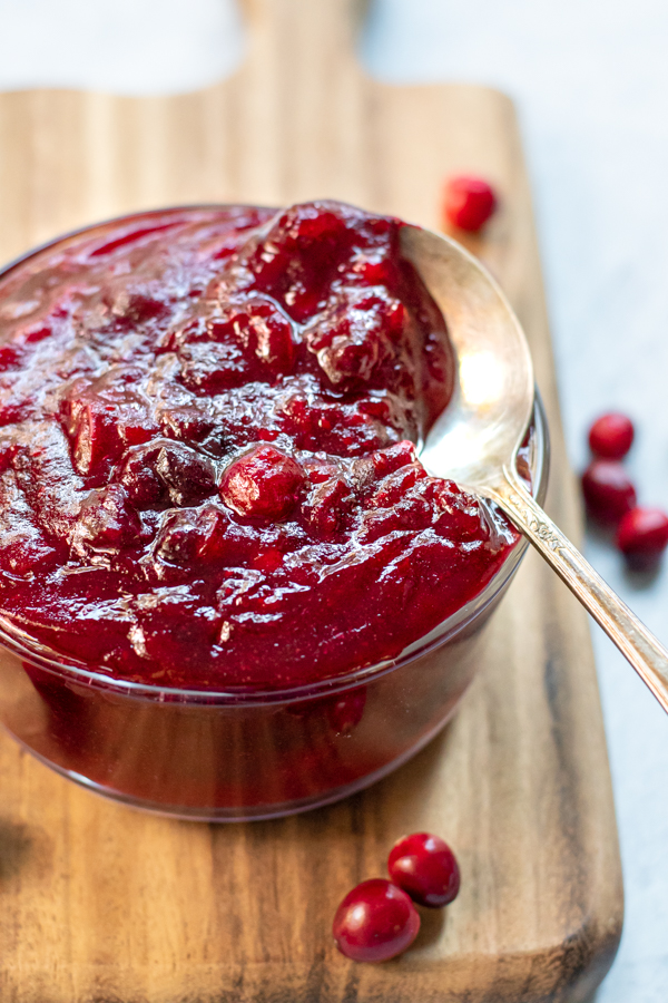 Small bowl of cranberry sauce with a spoon in the bowl sitting on a wooden cutting board next to loose fresh cranberries.