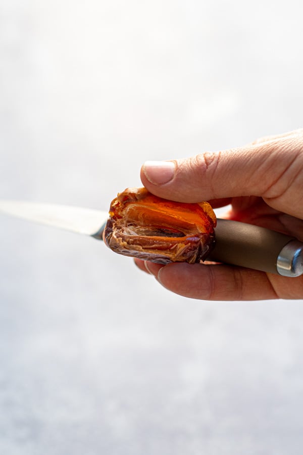A hand holding a paring knife and a sliced open date with the pit removed.