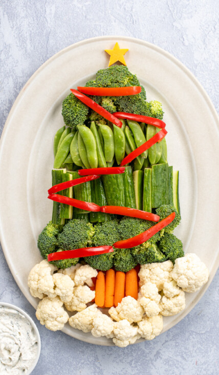 A platter containing a Christmas Tree Veggie Tray, green veggies are shaped to look like a tree with red bell pepper slices as decorations.