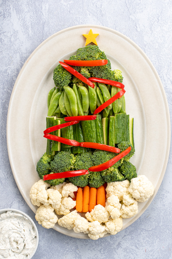 A platter containing a Christmas Tree Veggie Tray, green veggies are shaped to look like a tree with red bell pepper slices as decorations.