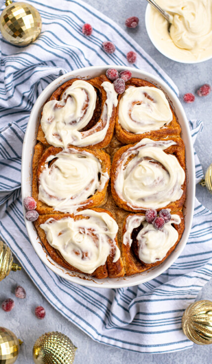 Overhead view of a white baking dish full of gingerbread cinnamon rolls topped with cream cheese frosting and garnished with sugared cranberries next to gold Christmas ornaments.